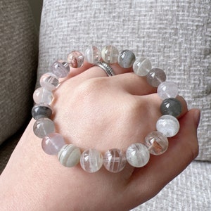 Peut inclure: Un bracelet composé de perles de quartz clair avec des inclusions grises, blanches et roses. Les perles sont rondes et ont une finition lisse et polie.