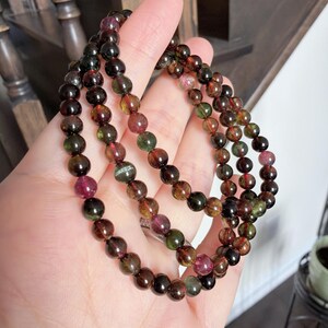 Puede incluir: Una pulsera de múltiples hilos con cuentas redondas pulidas en tonos verdes, marrones y rosas. Las cuentas están ensartadas, creando un efecto en capas. La pulsera se sostiene en una mano.