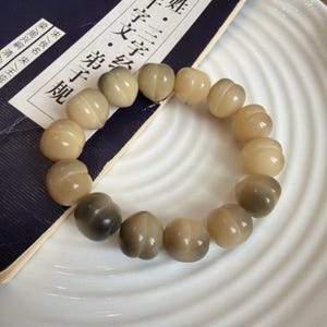 May include: A beaded bracelet composed of light brown, rounded beads. The beads have a slightly textured surface and are strung together to form a circular shape. The bracelet is resting on a white plate next to a book with Chinese characters.