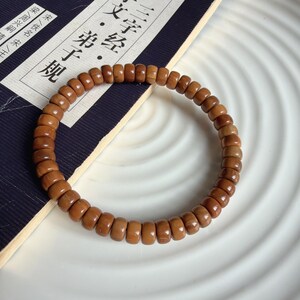 May include: A brown beaded bracelet made of small, cylindrical beads. The bracelet is circular and sits on a white, ridged surface. The beads have a smooth, polished appearance. A book with Chinese characters is partially visible in the background.