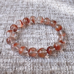 Könnte beinhalten: Ein Armband aus klaren Quarzperlen mit roten und orangefarbenen Einschlüssen. Die Perlen sind rund und haben eine glatte, polierte Oberfläche.