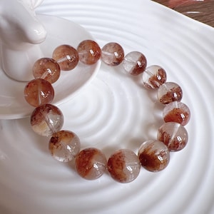 Puede incluir: Un brazalete hecho de cuentas de cuarzo transparente con inclusiones rojas y marrones. Las cuentas son redondas y tienen un acabado liso y pulido.