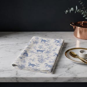 May include: A folded rectangular fabric napkin featuring a blue and white toile pattern with illustrations of cats interspersed among floral motifs. The fabric has a cream-colored base with a repeating design of various feline poses and foliage.