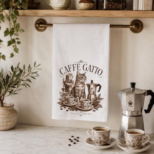 May include: A white kitchen towel featuring a brown graphic print of a cat sitting next to a moka pot and two cups of coffee. The text CAFFE GATTO and ITALIAN ESPRESSO is printed on the towel. The design includes a small building illustration.