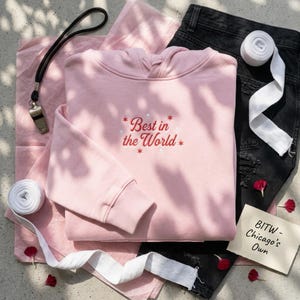 May include: A light pink hoodie with the embroidered text "Best in the World" in red and white. The hoodie is laid out with black jeans, a whistle, and white tape. The phrase "BITW - Chicago's Own" is on a small card.