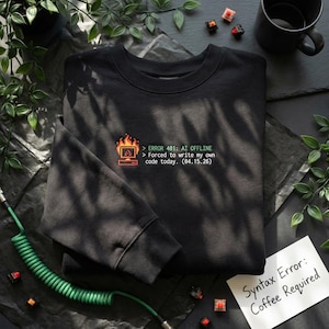 May include: Black crew neck sweatshirt featuring a computer error graphic and the text "ERROR 401: AI OFFLINE". A small note reads "Syntax Error: Coffee Required". The sweatshirt is a dark colour.