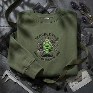 May include: Olive green sweatshirt with the text "SCOURGE FILE" and a graphic of a vial with a biohazard symbol, a planet, and a mustache. The text "DON'T LEAVE ME HANGING HERE" is also present. A note reads "Specimen 04: Do not break seal."