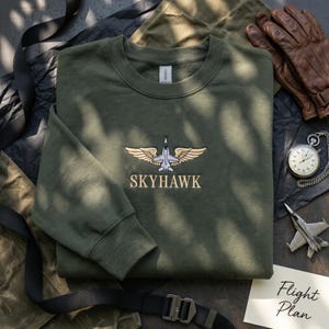 Op de afbeelding: Een olijfgroene sweatshirt met het woord "SKYHAWK" geborduurd onder een gestileerd vliegtuig met vleugels. Het ontwerp is beige en grijs. De sweatshirt ligt op een donkere ondergrond met een zwarte band en een klein notitieblok met "Flight Plan".