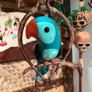 May include: A hand-carved, wooden parrot figurine with a vibrant teal body, red beak, and black eye. The bird is perched on a brown branch within a decorative wooden frame, showcasing a tropical theme. The background features Polynesian-inspired decor.