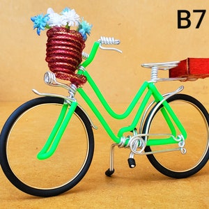 May include: A handcrafted decorative bicycle sculpture. The bicycle features a bright green frame, black tires, and a basket filled with blue and white flowers. The seat is a small red block. The text "B7" is visible.