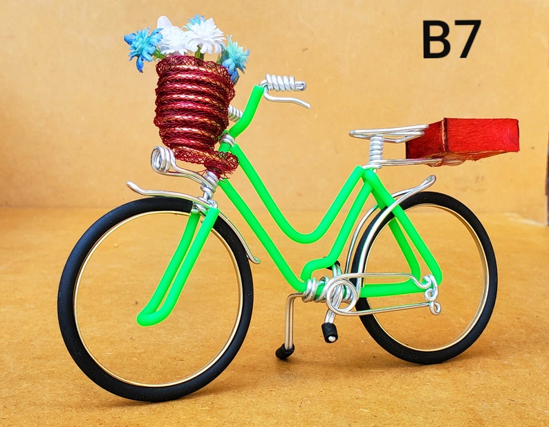May include: A handcrafted decorative bicycle sculpture. The bicycle features a bright green frame, black tires, and a basket filled with blue and white flowers. The seat is a small red block. The text "B7" is visible.