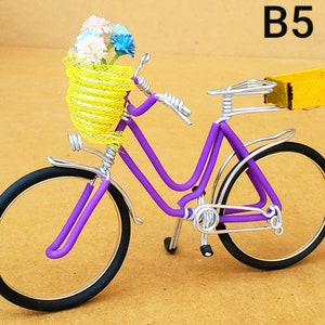 May include: A miniature purple bicycle with black tires and silver accents. The bicycle has a yellow wire basket filled with colorful flowers and a yellow wooden box on the back. The text "B5" is in the upper right corner.