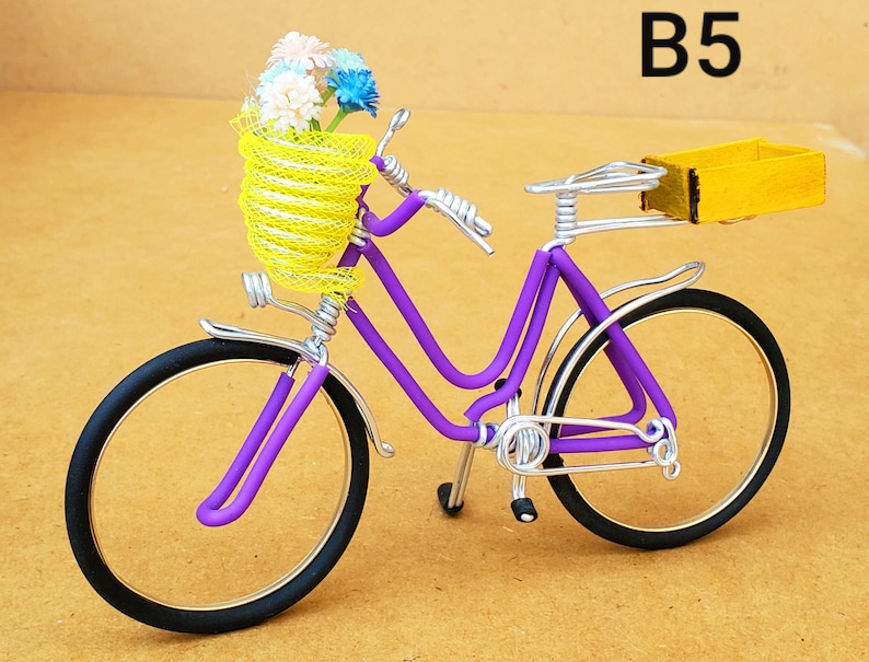 May include: A miniature purple bicycle with black tires and silver accents. The bicycle has a yellow wire basket filled with colorful flowers and a yellow wooden box on the back. The text "B5" is in the upper right corner.
