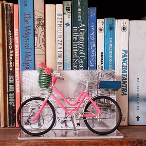 May include: A decorative pink bicycle figurine with black wheels and a small blue box on the back. The bicycle is in front of a black and white photo and a row of books. The bicycle has a basket with pink flowers.