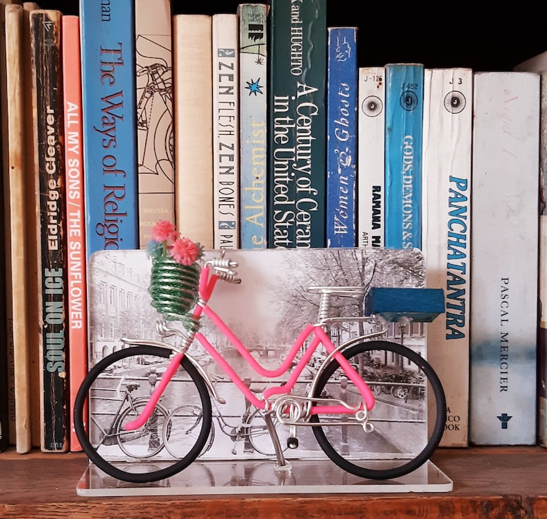 May include: A decorative pink bicycle figurine with black wheels and a small blue box on the back. The bicycle is in front of a black and white photo and a row of books. The bicycle has a basket with pink flowers.