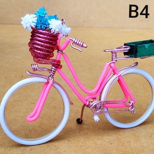 May include: A pink wire bicycle with white tires and a basket filled with blue and white flowers. A small green box is attached to the back. The text "B4" is visible in the upper right corner.