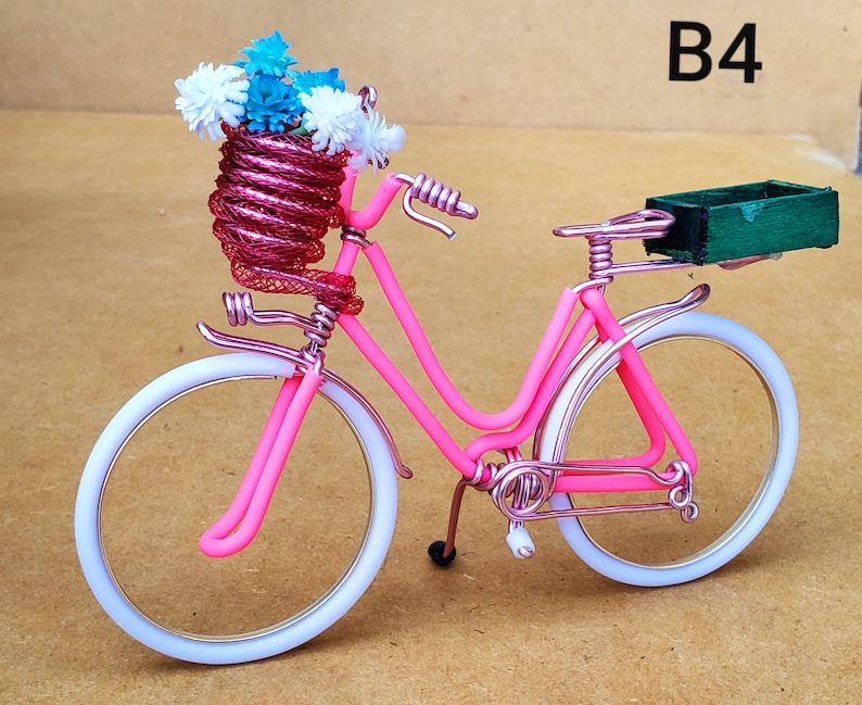 May include: A pink wire bicycle with white tires and a basket filled with blue and white flowers. A small green box is attached to the back. The text "B4" is visible in the upper right corner.