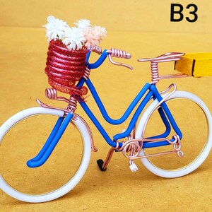 May include: A miniature bicycle sculpture with a blue frame, white tires, and copper-colored accents. The front basket is filled with white and pink flowers. A small yellow box is attached to the back. The text "B3" is in the upper right corner.