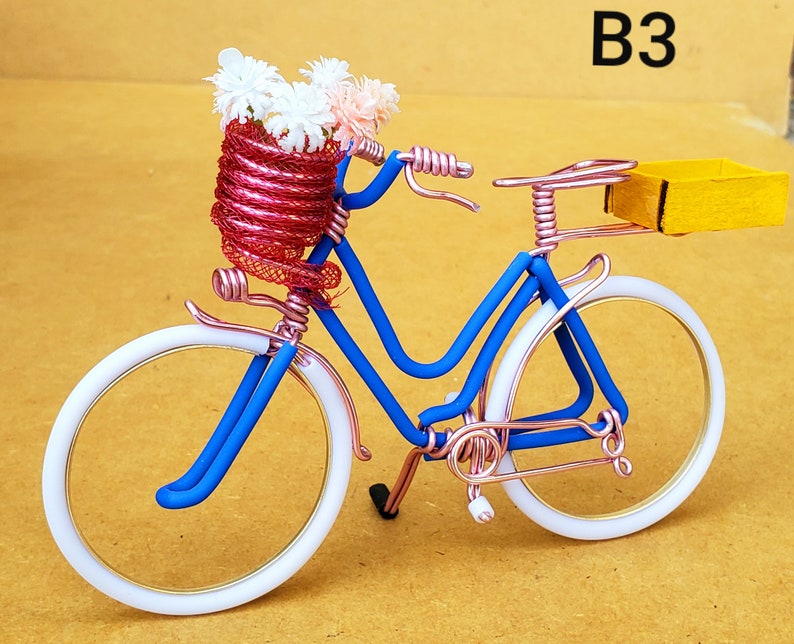 May include: A miniature bicycle sculpture with a blue frame, white tires, and copper-colored accents. The front basket is filled with white and pink flowers. A small yellow box is attached to the back. The text "B3" is in the upper right corner.
