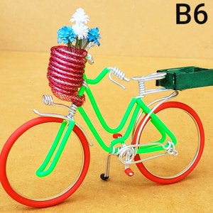 May include: A decorative bicycle sculpture made of wire, featuring a green frame, red wheels, and a basket filled with blue and white flowers. The sculpture has a green box on the back and the text "B6" in the upper right corner.