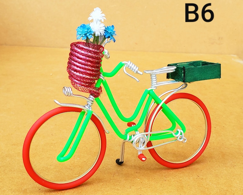 May include: A decorative bicycle sculpture made of wire, featuring a green frame, red wheels, and a basket filled with blue and white flowers. The sculpture has a green box on the back and the text "B6" in the upper right corner.