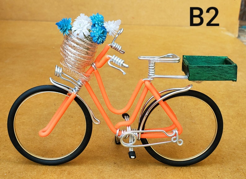 May include: An orange wire bicycle sculpture with black tires and gold rims. The front basket holds blue and white flowers. A green wooden box is attached to the back. The text "B2" is in the upper right corner.