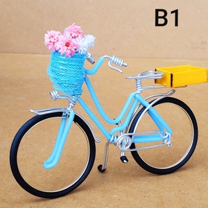 May include: A light blue decorative bicycle with black tires and silver wire accents. The front basket holds pink and white flowers. A yellow box is attached to the back. The text "B1" is visible in the upper right corner.