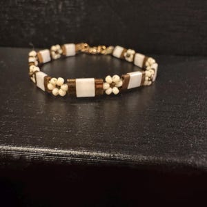 以下が含まれることがあります： 白と金のビーズで作られたブレスレットで、小さな白い花のアクセントが付けられています。