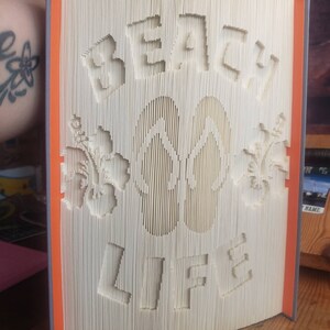 May include: A white book with the words "Beach Life" folded into the pages. The book also features two flip flops and two flowers.