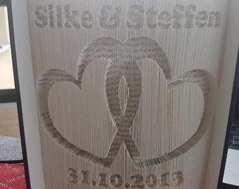 May include: A white book with the words "Silke & Stefan" and "31.10.2019" cut out of the pages. The book is folded into a three-dimensional heart shape.