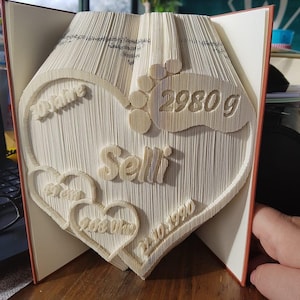 Könnte beinhalten: Ein weißes Buch, das zu einem Herz gefaltet ist, mit dem Namen "Selin" und dem Datum "20.06.1990" im Inneren. Das Herz ist von zwei kleineren Herzen mit dem Text "2980g" und "30.06.1990" umgeben.