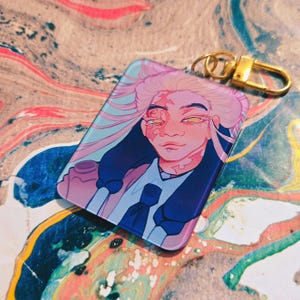 Puede incluir: Un llavero rectangular con un retrato estilizado de una persona con cabello rosado y ropa azul. El retrato tiene una est&eacute;tica suave y colorida con herrajes dorados. El fondo es un remolino de colores.