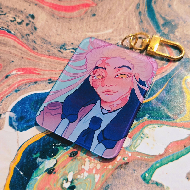 Puede incluir: Un llavero rectangular con un retrato estilizado de una persona con cabello rosado y ropa azul. El retrato tiene una est&eacute;tica suave y colorida con herrajes dorados. El fondo es un remolino de colores.