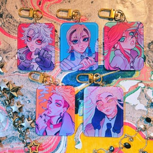 Puede incluir: Una colecci&oacute;n de llaveros rectangulares con retratos coloridos de estilo anime y cierres dorados. Cada llavero muestra un personaje diferente con peinados y expresiones &uacute;nicas, sobre fondos vibrantes.