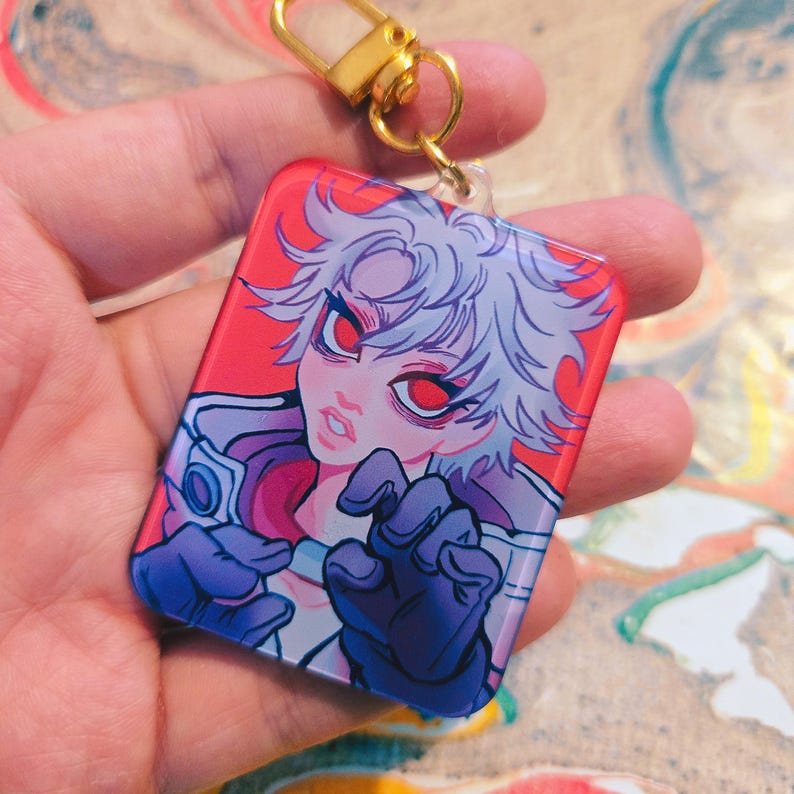 Puede incluir: Un llavero rectangular con un personaje de estilo anime con cabello blanco y ojos rojos. El personaje est&aacute; sobre un fondo rojo. El llavero tiene un cierre dorado.
