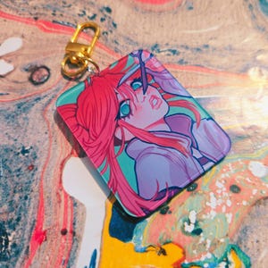 Puede incluir: Un llavero rectangular con una ilustraci&oacute;n de estilo anime de un personaje con cabello rosado y ojos turquesa. El llavero tiene un cierre dorado. La obra de arte est&aacute; sobre un fondo turquesa y morado.