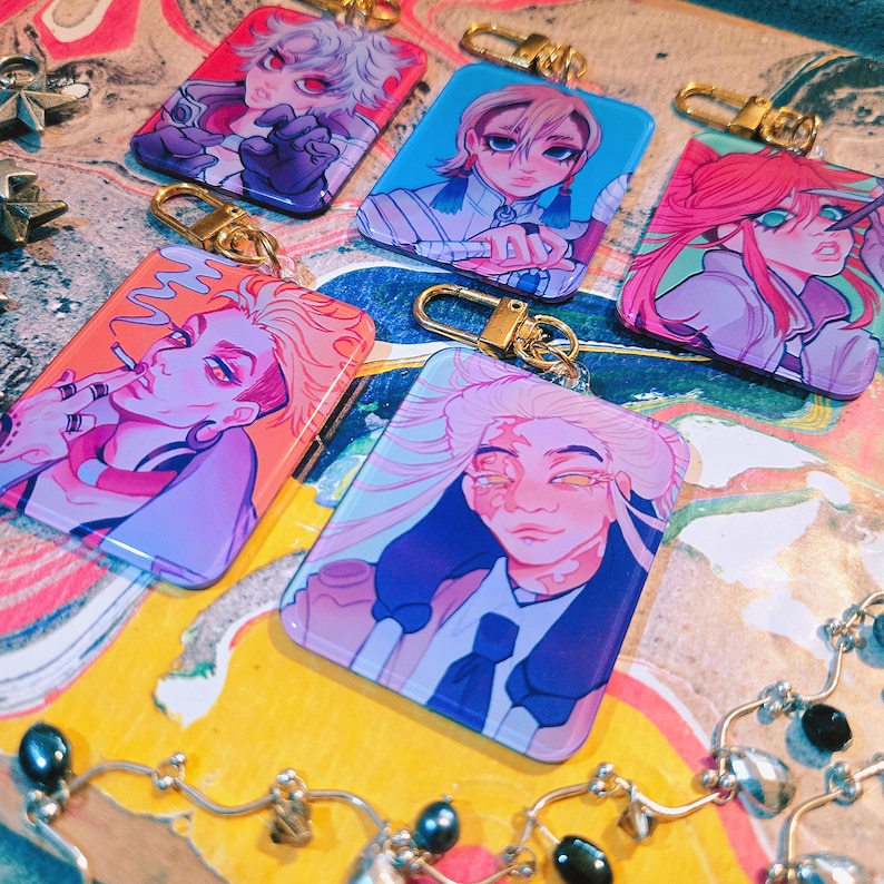 Puede incluir: Una colecci&oacute;n de llaveros rectangulares con ilustraciones coloridas de personajes de estilo anime. Cada llavero tiene un cierre dorado. Los personajes tienen varios colores de pelo y expresiones, sobre fondos vibrantes de color naranja, azul y rosa.