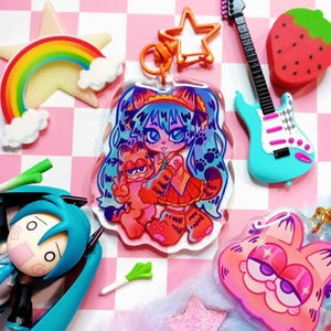 May include: A clear acrylic charm featuring a cartoon character with blue hair and a red and white outfit holding a cartoon orange cat. The charm is attached to a silver chain with a star-shaped charm.