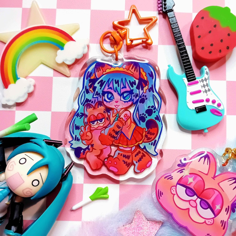 May include: A clear acrylic charm featuring a cartoon character with blue hair and a red and white outfit holding a cartoon orange cat. The charm is attached to a silver chain with a star-shaped charm.