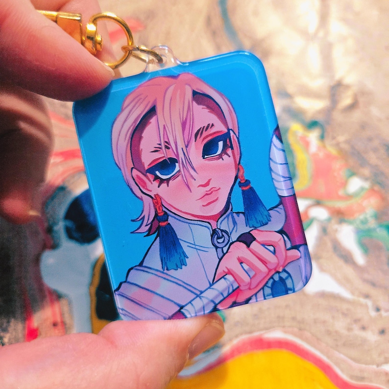 Puede incluir: Un llavero rectangular azul con una ilustraci&oacute;n de estilo anime de una persona con cabello rosado, ojos azules y aretes de borlas azules. El llavero tiene un cierre dorado.
