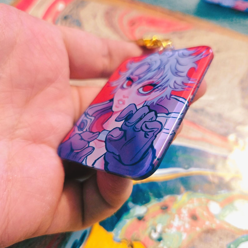 Puede incluir: Colgante rectangular con un personaje de anime estilizado con cabello blanco y ojos rojos sobre un fondo rojo. El personaje tiene una mano enguantada. El colgante tiene un lazo dorado en la parte superior.