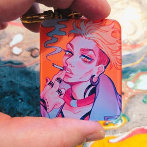 Puede incluir: Un llavero rectangular con una vibrante ilustraci&oacute;n de una persona fumando. La obra de arte presenta a una persona con cabello naranja, que lleva una chaqueta azul y varios anillos. El fondo es naranja y el llavero tiene un cierre dorado.