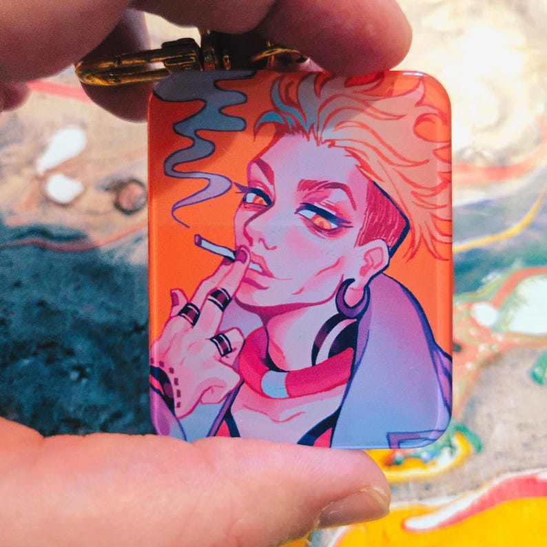 Puede incluir: Un llavero rectangular con una vibrante ilustraci&oacute;n de una persona fumando. La obra de arte presenta a una persona con cabello naranja, que lleva una chaqueta azul y varios anillos. El fondo es naranja y el llavero tiene un cierre dorado.