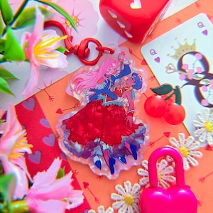 Può includere: Un portachiavi in acrilico trasparente con un'illustrazione colorata di due figure in un abbraccio romantico. Il portachiavi ha un fermaglio rosso. Altri oggetti includono un dado rosso a forma di cuore, una carta da gioco e un lucchetto rosa a forma di cuore.
