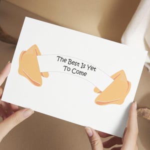 Könnte beinhalten: Eine weiße Karte mit einer Illustration eines Glückskekses und dem Text "The Best Is Yet To Come". Der Glückskeks ist hellorange mit einer dunkelorangen Umrandung. Die Karte wird von zwei Händen gehalten.