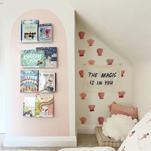May include: A pink wall with a white shelf unit holding a variety of children's books. The shelf unit is decorated with a pink arch. The wall is decorated with pink seashell stickers and a white banner with the text "The Magic Is In You".