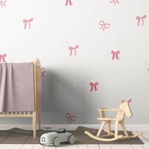 May include: A nursery scene with a light wood crib, a rocking horse, and a toy car. The wall is decorated with pink bow decals. A pink blanket is draped over the crib. The floor has a brown rug.