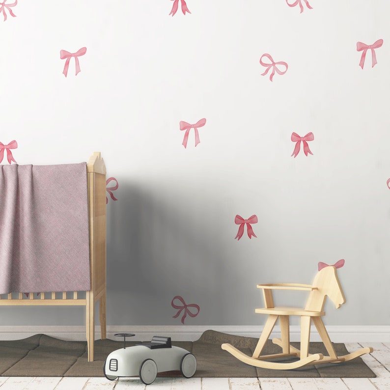 May include: A nursery scene with a light wood crib, a rocking horse, and a toy car. The wall is decorated with pink bow decals. A pink blanket is draped over the crib. The floor has a brown rug.