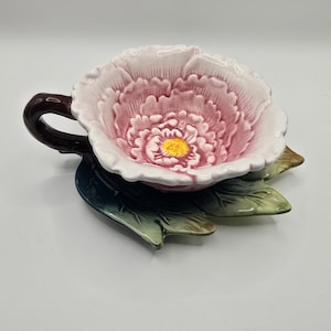 Puede incluir: Un juego de taza y platillo de cerámica con forma de flor de peonía. La taza es rosa y blanca con un centro amarillo, y el asa es marrón oscuro. El platillo es verde con detalles marrones, que se asemejan a hojas. El juego está diseñado para parecerse a una flor.