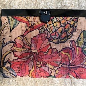 May include: A cork clutch with a floral print featuring red, yellow, and green hibiscus flowers and a pineapple. The clutch has a black metal clasp.